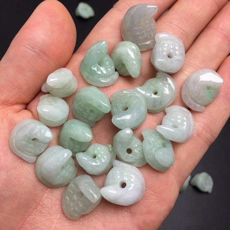 Pode incluir: Uma cole&ccedil;&atilde;o de pequenos pingentes de jade esculpidos em v&aacute;rias formas, incluindo p&aacute;ssaros e formas abstratas. Os pingentes s&atilde;o principalmente verde-claro e brancos, com um acabamento liso e polido. Os pingentes s&atilde;o segurados em uma m&atilde;o.
