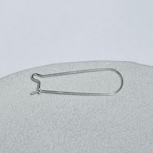 Könnte beinhalten: Ein silberfarbener Ohrringdraht in Form einer länglichen, abgerundeten Schlaufe mit einem kleinen Hakenverschluss an einem Ende, der zur Befestigung von Schmuckkomponenten in einer einfachen, funktionalen Drahtkonstruktion dient.