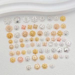 Könnte beinhalten: Eine Sammlung von blütenförmigen Metallperlenkappen in verschiedenen Größen und Ausführungen. Die Kappen sind in Gold-, Silber- und Roségoldtönen gehalten und weisen aufwendige Designs und Texturen auf. Diese dekorativen Komponenten sind ideal für die Schmuckherstellung und Bastelprojekte.