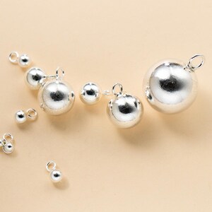May include: A collection of silver-colored spherical charms of varying sizes, each featuring a small circular loop at the top for attachment. The metallic spheres have a smooth, reflective surface and are arranged in a scattered formation.
