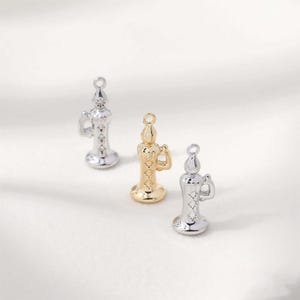 May include: Three small candle-shaped charms in silver and gold tones. Each charm has a loop at the top for hanging, a handle, and star-shaped cutouts. The charms are on a white background.