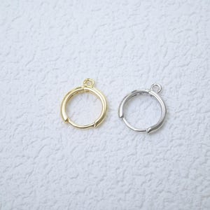 May include: Two small hoop earrings, one gold-toned and one silver-toned, are displayed against a textured white background. Each earring has a circular design with a small loop at the top for attachment. The earrings are simple and elegant.