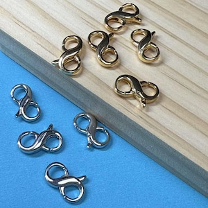 May include: A close-up of eight gold and silver infinity lobster clasps. The clasps are arranged in two rows on a wooden surface. The clasps are a popular jewelry finding for making necklaces, bracelets, and other jewelry.
