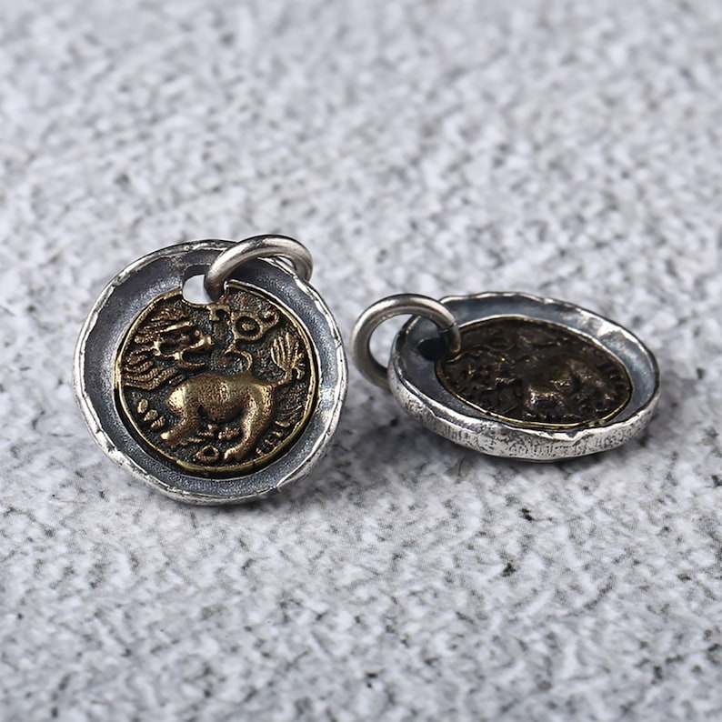 May include: Two silver and gold tone pendants featuring a lion design. The pendants have a textured edge and a loop for attaching to a chain.