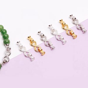 May include: A collection of infinity-shaped jewelry clasps in gold and silver tones. The clasps are displayed on a white and purple background, with a green beaded bracelet visible on the left side. These clasps are designed for jewelry making.