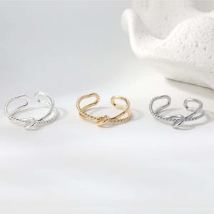 May include: Three adjustable rings in silver and gold tones. Each ring features a twisted rope design on the band and a double-loop design. The rings are displayed on a white surface.