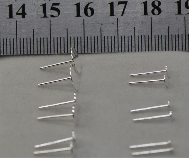 Peut inclure: Gros plan sur des tiges d'oreille en argent avec des sommets ronds plats. Les tiges sont dispos&eacute;es en rang&eacute;es sur une surface blanche. Une r&egrave;gle est visible en arri&egrave;re-plan.