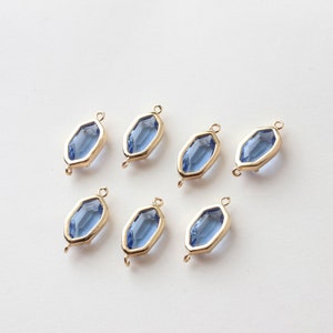 May include: Seven gold-toned, geometric charms with a blue-colored, faceted glass center. Each charm has a loop at the top and bottom for attaching to jewelry. The charms are arranged on a white surface.