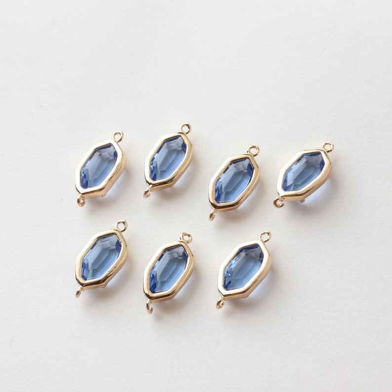 May include: Seven gold-toned, geometric charms with a blue-colored, faceted glass center. Each charm has a loop at the top and bottom for attaching to jewelry. The charms are arranged on a white surface.