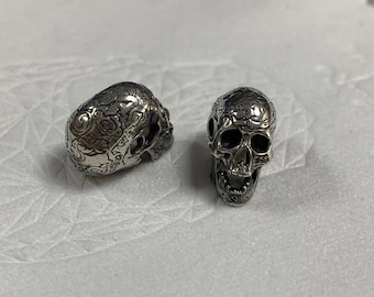 May include: Two silver skull-shaped charms, one with a detailed floral design and the other with a more traditional skull design.