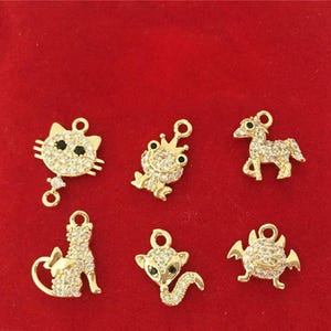 May include: A collection of six gold-toned metal charms featuring animal and creature shapes, including a cat face, a crowned frog, a horse, a sitting cat, a fox, and a winged creature, all encrusted with clear and black cubic zirconia stones.