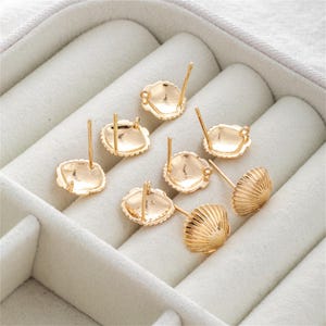 Pode incluir: Brincos de botão dourados em forma de concha. Os brincos têm um design detalhado e canelado na concha e um pino para perfuração. Os brincos são exibidos em uma superfície branca texturizada.