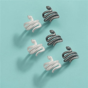 May include: Six snake-shaped rings in silver and dark gray metal. The rings are coiled, with the snake's head at the top. The rings are displayed on a light blue surface.