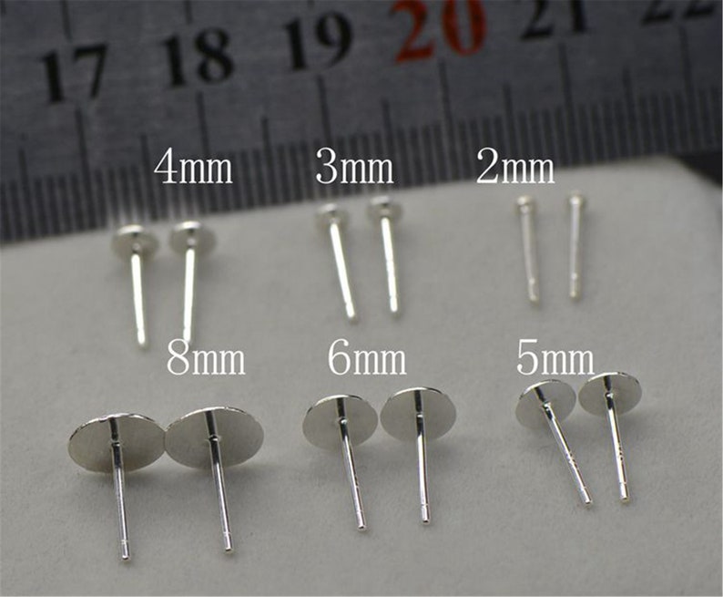 Peut inclure: Assortiment de tiges d'oreilles en argent de diff&eacute;rentes tailles, notamment 2 mm, 3 mm, 4 mm, 5 mm, 6 mm et 8 mm. Les tiges d'oreilles sont pr&eacute;sent&eacute;es sur une surface blanche avec une r&egrave;gle en arri&egrave;re-plan.