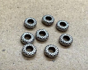 May include: Eight silver-coloured beads with a patterned design. Each bead is a small, hollow cylinder with a detailed, textured surface. The beads are arranged on a neutral, light brown surface.