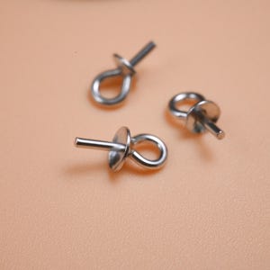 Pode incluir: Três pinos de olho prateados com um laço e um disco plano. Os pinos estão dispostos sobre uma superfície cor de pêssego. Estes achados são usados na fabricação de joias para conectar contas e outros componentes.