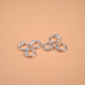May include: Six small, silver-colored, circular jewelry components are arranged on a peach-colored surface. Each component has a smooth, rounded shape with a slight indentation on one side. The components appear to be made of metal.
