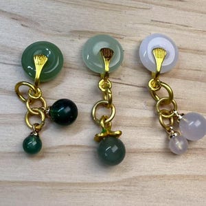 May include: Three pairs of dangle earrings with gold-coloured metal accents. Each earring features a circular jade stone at the top, with dangling jade and gemstone beads. The earrings vary in colour, including green and white.