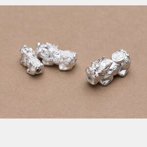 Op de afbeelding: Drie zilveren Chinese drakencharms met ingewikkelde details en muntachtige accenten. De charms liggen op een bruine ondergrond.