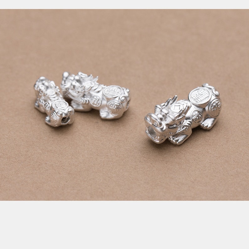 Op de afbeelding: Drie zilveren Chinese drakencharms met ingewikkelde details en muntachtige accenten. De charms liggen op een bruine ondergrond.