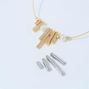 Può includere: Una collana dorata con un design lineare, caratterizzata da barre verticali e perle finte. Ulteriori componenti di gioielleria argentati a forma di barra sono sparsi sotto. La collana ha una delicata catena a filo.