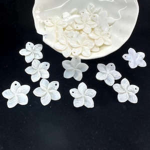 Może przedstawiać: Białe, kwiatowe zawieszki z muszli o perłowym połysku. Zawieszki są rozrzucone na czarnej powierzchni i w białej miseczce w kształcie muszli. Każda zawieszka ma mały otwór do nawlekania.