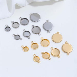May include: An assortment of jewelry making components in silver and gold tones. The components include round bezel settings and flat circles with loops for attaching to chains or other elements. The pieces vary in size and are arranged on a white surface.