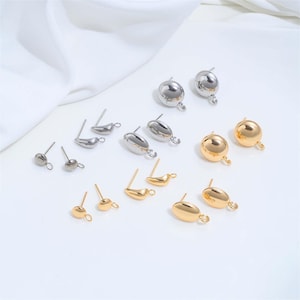 Op de afbeelding: Een verzameling metalen oorstekers in zilver- en goudtinten, met diverse gladde, ronde en ovale vormen. Elk oorbellelement is voorzien van een klein oogje aan de basis, ontworpen voor het bevestigen van bedels aan de sieraden.