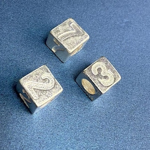 Può includere: Tre dadi color argento con i numeri incisi 2, 3 e 7. I dadi sono di forma quadrata e hanno un aspetto leggermente usurato e vintage. Lo sfondo è di colore blu.