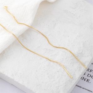 Może przedstawiać: Złote kolczyki z nitką. Kolczyki mają delikatny łańcuszek z małym pręcikiem na końcu. Kolczyki są prezentowane na białej powierzchni.