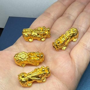 Puede incluir: Cuatro amuletos de león de color dorado, cada uno con detalles intrincados y un diseño redondo similar a una moneda en la parte posterior. Los amuletos se muestran en una mano.