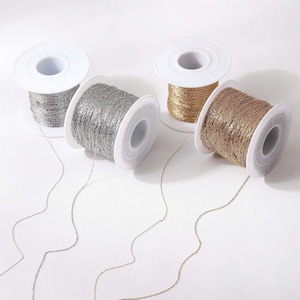 Puede incluir: Cuatro carretes de cadena metálica en tonos plateado, dorado y bronce. La cadena está enrollada alrededor de carretes de plástico blanco. Las cadenas están desenrolladas, creando un efecto delicado y fluido. Ideal para bisutería y manualidades.