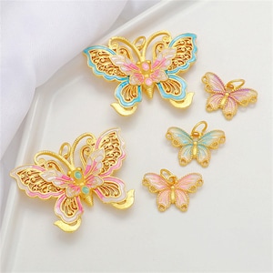 Puede incluir: Encantos de mariposa dorados con detalles de esmalte rosa, azul y blanco. Los encantos varían en tamaño y presentan intrincados diseños de filigrana. Las mariposas se muestran sobre una superficie blanca.