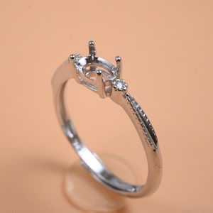 May include: A silver ring with an oval gemstone setting and a delicate band. The ring features a small, round accent stone near the setting and a split design on the band. The ring is displayed on a clear stand.