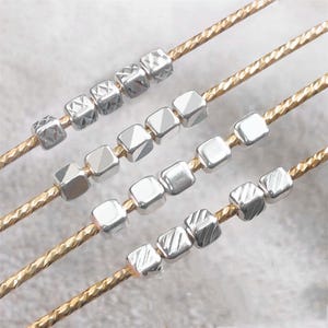 Può includere: Primo piano di quattro fili di gioielli. Ogni filo presenta perline quadrate color argento con vari motivi geometrici. Le perline sono infilate su un filo dorato strutturato. Lo sfondo è di un colore morbido e neutro.