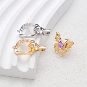 May include: Jewelry clasps and a flower-shaped charm. Two clasps are shown, one silver-toned and one gold-toned, both with a rectangular loop and a connecting piece. The flower charm is gold-toned with a purple gemstone center.