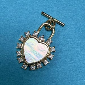 May include: A gold heart-shaped pendant with a white iridescent center and a clear crystal halo. The pendant is attached to a gold toggle clasp.