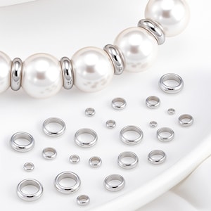 May include: A collection of smooth, polished silver-toned metal spacer beads in various sizes, arranged alongside a partial strand of large, round, white faux pearls separated by matching silver-toned metal spacer beads.