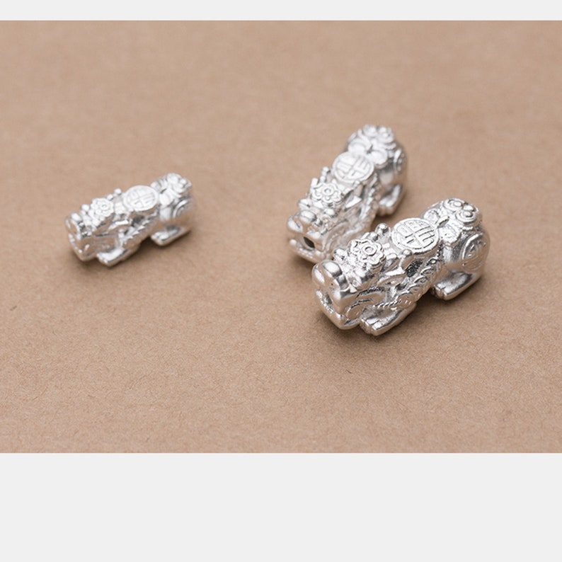 Op de afbeelding: Drie zilveren Chinese drakencharms met ingewikkelde details. De charms zijn in de vorm van een draak met een munt op zijn rug. De charms liggen op een bruine ondergrond.