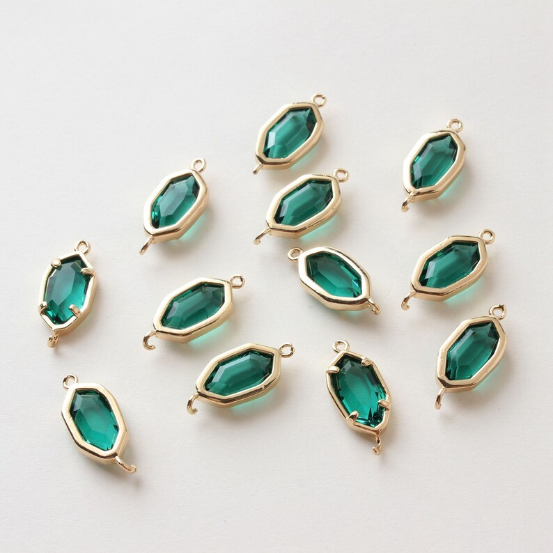 May include: A collection of emerald green gemstone pendants with gold-colored frames. Each pendant has a faceted, elongated hexagonal shape and a small loop at each end for attaching to jewelry. The pendants are arranged on a white surface.