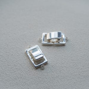 Puede incluir: Dos cierres rectangulares plateados para joyería. Cada cierre tiene un marco rectangular con una barra elevada articulada en el centro. Pequeños bucles están unidos al marco para conectar a la joyería. Los cierres están sobre un fondo neutro.