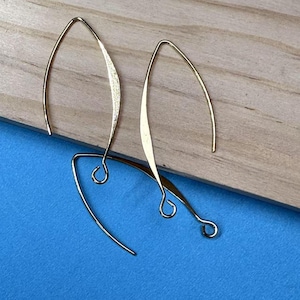Può includere: Orecchini pendenti dorati e delicati. Gli orecchini hanno un design curvo e allungato con un elemento testurizzato a forma di foglia. Sono esposti su una superficie di legno chiaro su sfondo blu.
