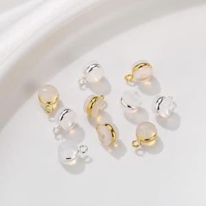 Puede incluir: Un conjunto de pequeños dijes redondos de color blanco translúcido, cada uno con una banda metálica central y una pequeña anilla circular en la parte superior. Algunos tienen bandas y anillas doradas, otros tienen bandas y anillas plateadas.