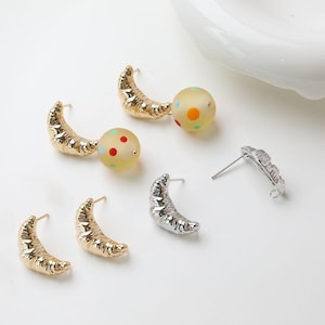 May include: A collection of earrings featuring a crescent moon design. Some earrings are gold-toned, while others are silver-toned. Some earrings have a round, translucent bead with colorful polka dots. The earrings are displayed on a white surface.