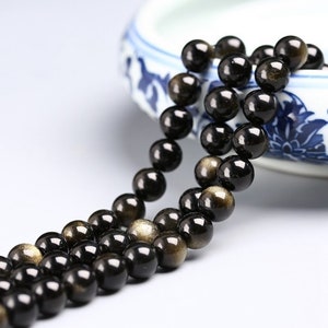 May include: A strand of black beads with gold flecks. The beads are round and smooth, and they are strung on a clear thread. The beads are arranged in a loose, flowing pattern.