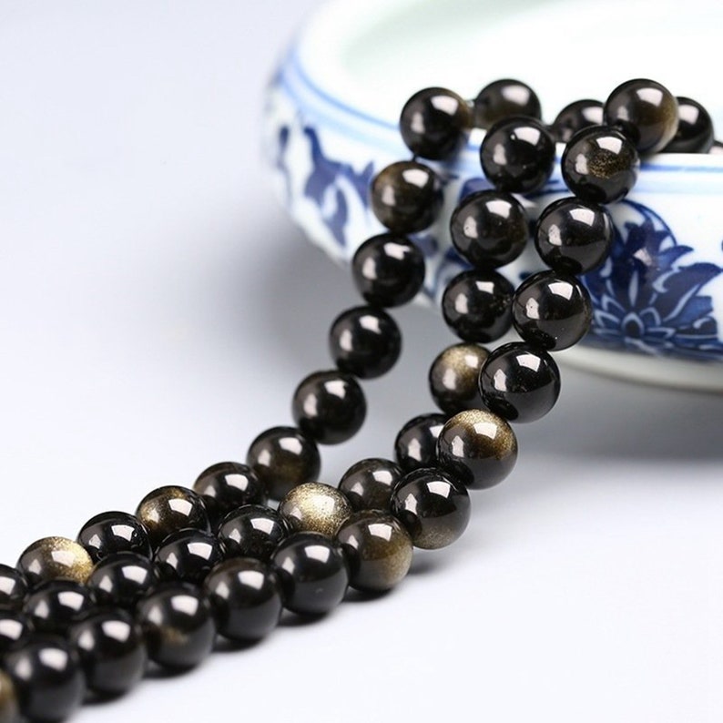 May include: A strand of black beads with gold flecks. The beads are round and smooth, and they are strung on a clear thread. The beads are arranged in a loose, flowing pattern.