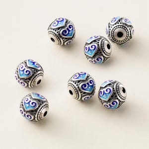 Può includere: Sei perline sferiche caratterizzate da una struttura in filigrana di metallo color argento con motivi a cuore in smalto blu intarsiato. Ogni perlina presenta un foro centrale e un bordo testurizzato attorno all'apertura, con un motivo metallico dettagliato.