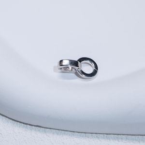 Puede incluir: Un pequeño cierre de joyería plateado con un diseño circular. El cierre tiene una sección con bisagras y un componente en forma de anillo, probablemente para sujetarlo a un collar o pulsera. El cierre está sobre una superficie blanca.