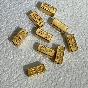 May include: A collection of eight rectangular gold-colored beads, each featuring a textured surface with two distinct circular stamped designs. The beads are arranged in a scattered pattern, showcasing a metallic finish and a small hole on each end.