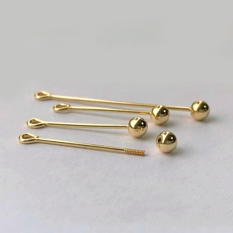 Peut inclure: Plusieurs &eacute;pingles de cravate et clous d'oreilles dor&eacute;s sont dispos&eacute;s sur une surface blanche. Les &eacute;pingles de cravate ont une boucle &agrave; une extr&eacute;mit&eacute; et une boule &agrave; l'autre. Les clous d'oreilles ont une boule et une tige filet&eacute;e.
